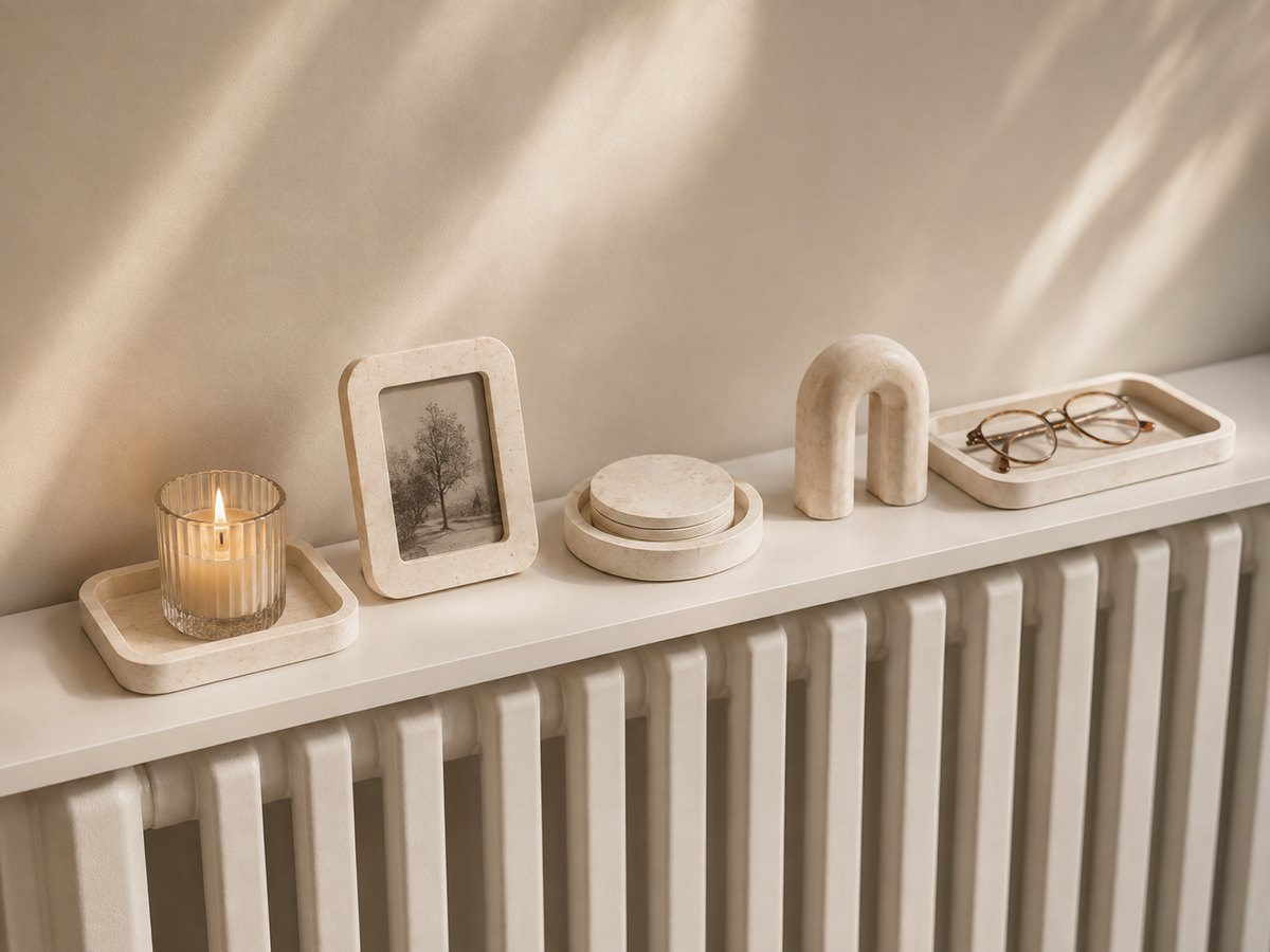A candle, photo frame, coasters, arch ornament and a tray with glasses styled along a white shelf above a radiator, in soft afternoon light