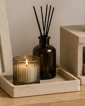 A lit candle in a ribbed glass holder beside an amber reed diffuser on a stone-effect tray