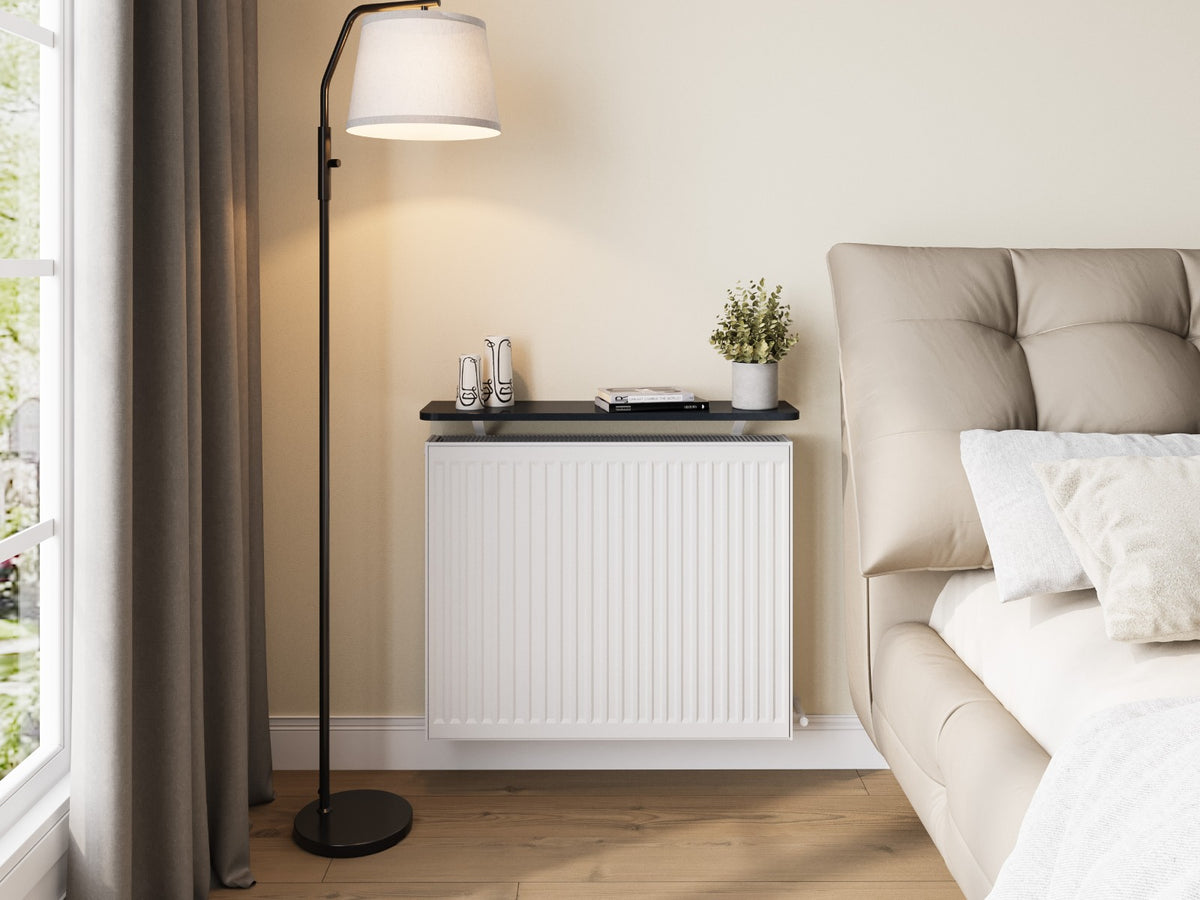 A bedroom with a black shelf over a white radiator, a bed close beside it and a floor lamp in front of the curtains
